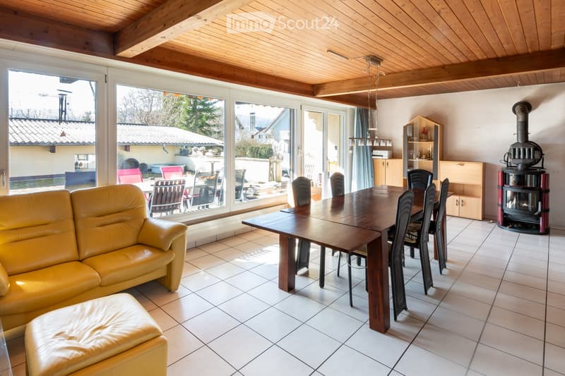 Bright living and dining area with a yellow leather sofa, ottoman, wooden dining table, chairs, and a fireplace. Large windows provide a view of the outdoor patio.