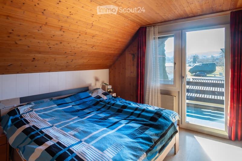 Cozy bedroom with wooden slanted ceiling, large window with red curtains, blue and black checkered bedspread, nightstand with lamp.