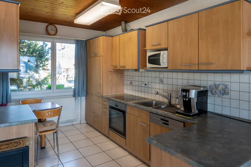 Well-lit kitchen with wooden cabinets, stainless steel appliances, and a tiled backsplash. Features a microwave, oven, sink, coffee maker, and a small dining area with a table and chair.