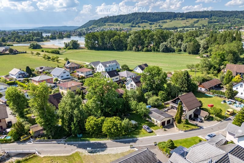 Aerial view of a residential neighborhood with multiple houses, surrounded by green fields, trees, and a river in the background.