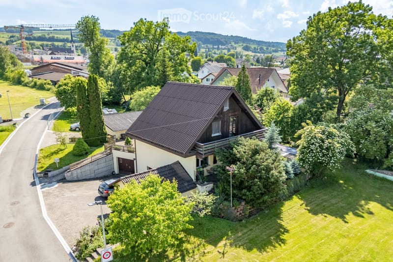 A two-story house with a dark brown roof and light yellow walls, surrounded by a well-maintained garden and trees. The house features a garage, a small balcony, and is situated in a residential area with neighboring houses and a scenic countryside view in