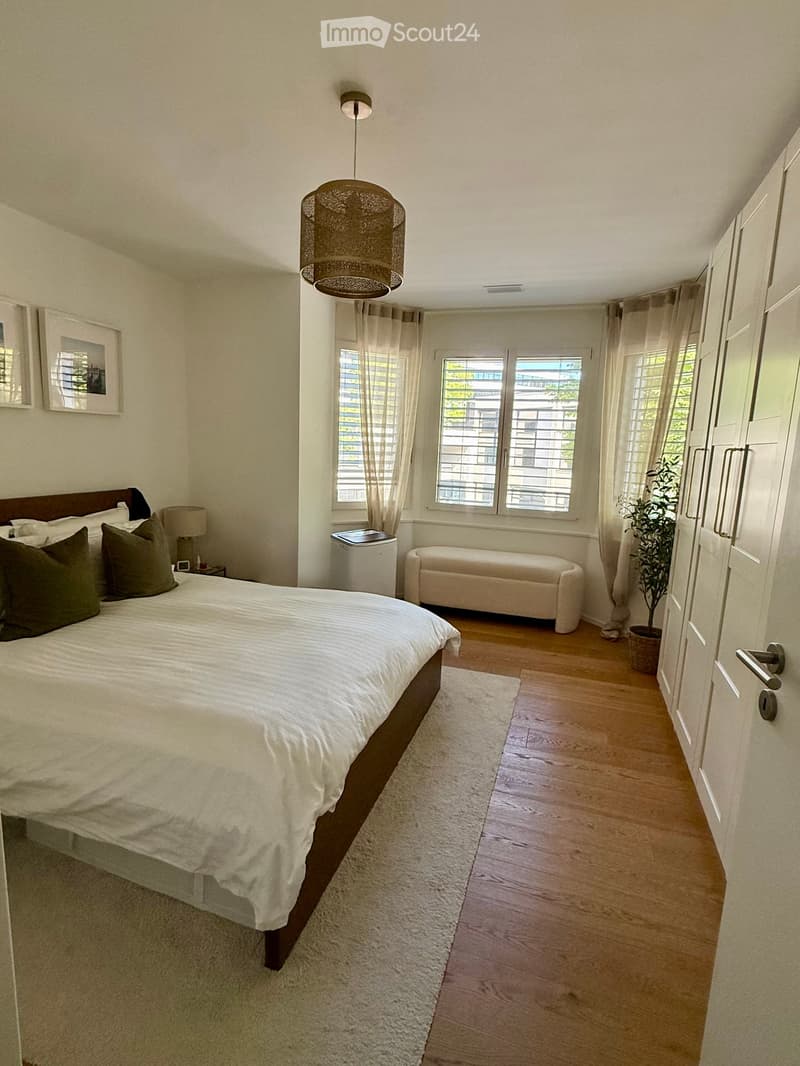 Well-lit bedroom with a large bed, nightstand, lamp, and a small bench near the window. Wooden flooring and white walls.