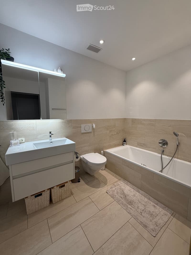 Modern bathroom with a large sink, toilet, bathtub, and a large mirror with built-in lighting. Beige tiles and a light-colored floor.