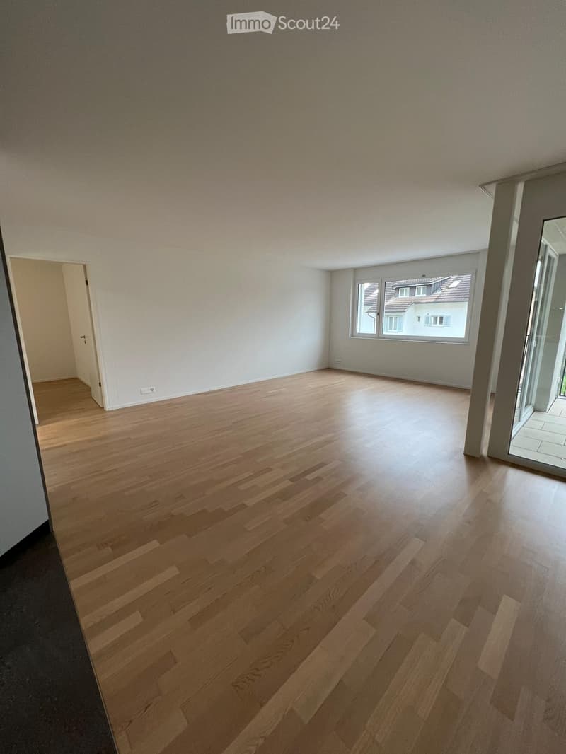 Empty room with light wooden flooring, white walls, and large windows offering a view of neighboring houses.