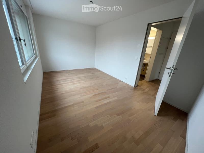 Empty room with light wooden flooring and white walls, featuring a large window and an open doorway leading to a small bathroom.