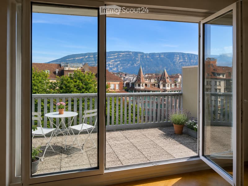 Balcony with a view of city rooftops and mountains, featuring a small table, two chairs, and potted plants.