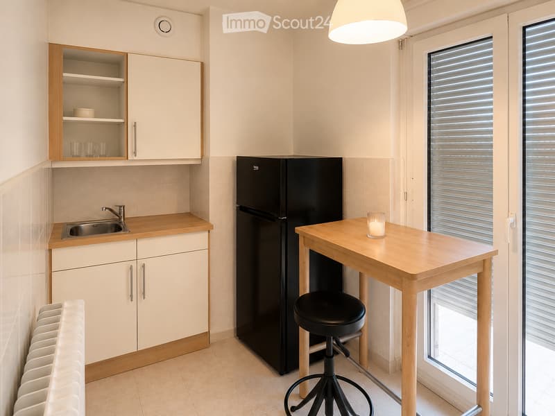 Modern kitchen with light wood cabinets, stainless steel sink, black refrigerator, wooden table with stool, and a candle on the table.