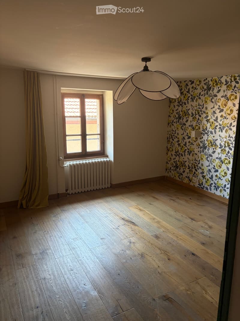 Empty room with wooden flooring, floral wallpaper, a window with a curtain, a radiator, and a modern pendant light fixture.