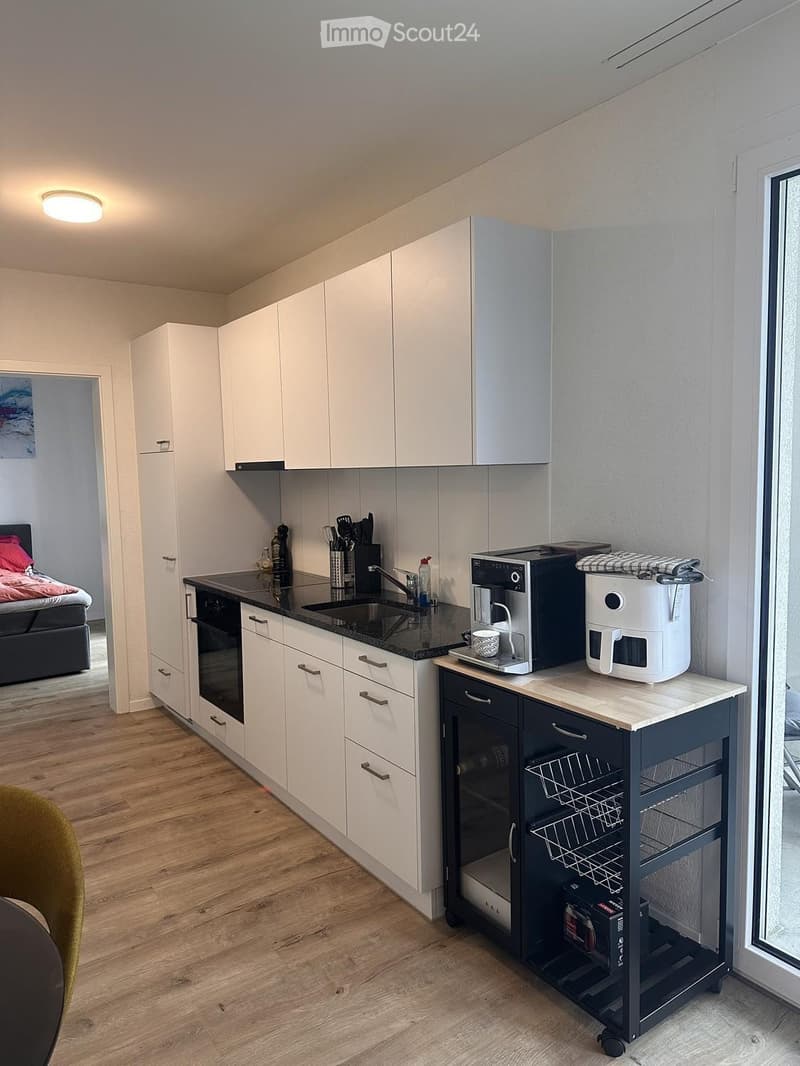 Modern kitchen with white cabinets, black countertop, and stainless steel appliances including a coffee maker and air fryer. A small island with a sink and a refrigerator is visible, along with a bed in the background.