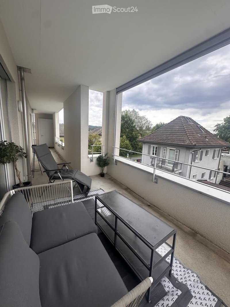 Balcony with a grey sofa, a black glass table, a wicker chair, and a lounge chair, overlooking a residential area with trees and houses.