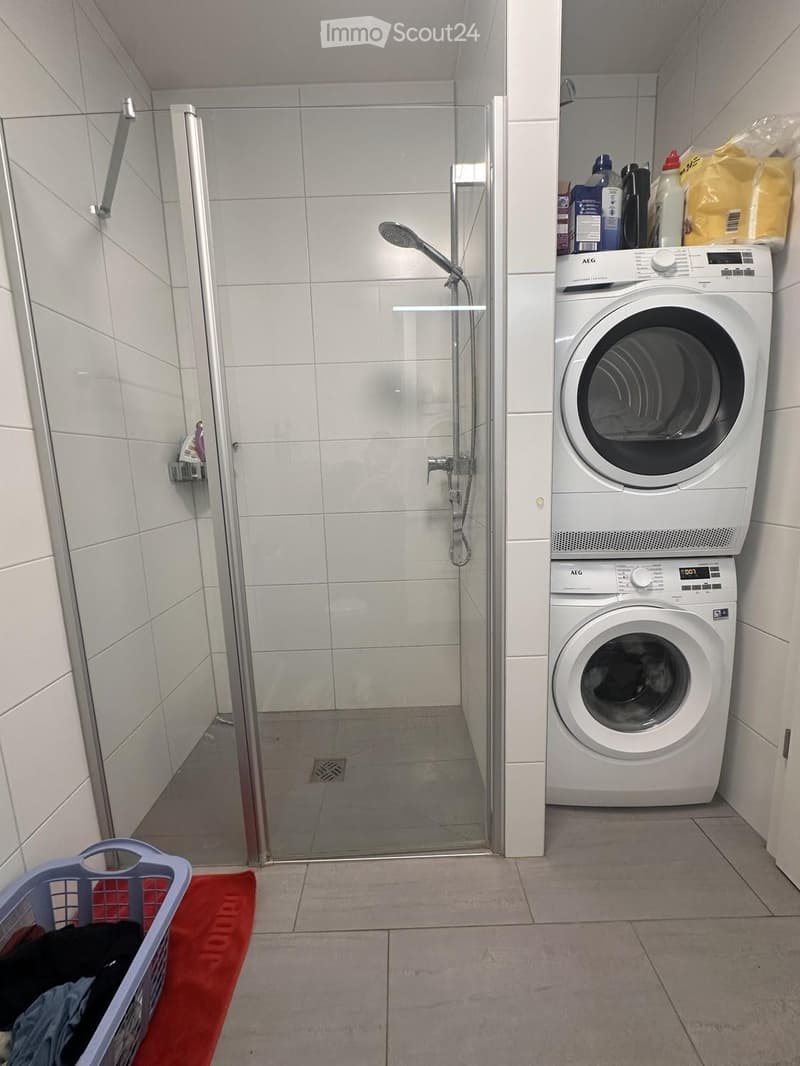 Modern bathroom with a glass-enclosed shower and a stacked washing machine and dryer unit. Laundry basket with clothes is visible on the floor.