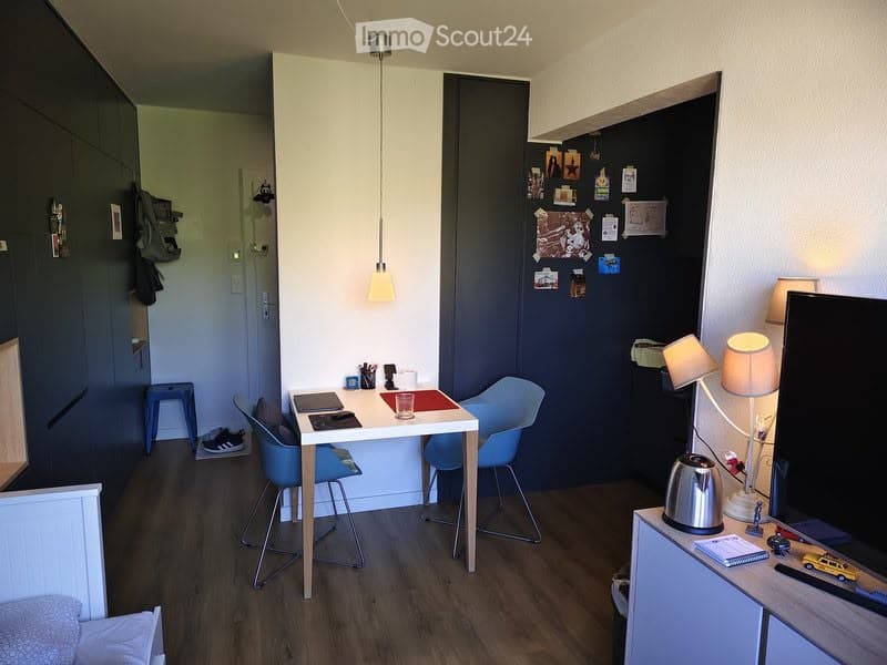A modern, compact room with a wooden floor, a small table with chairs, a stool, a lamp, a teapot, and a laptop on a side table. The walls are painted in dark tones with various decorations and a hanging light fixture.