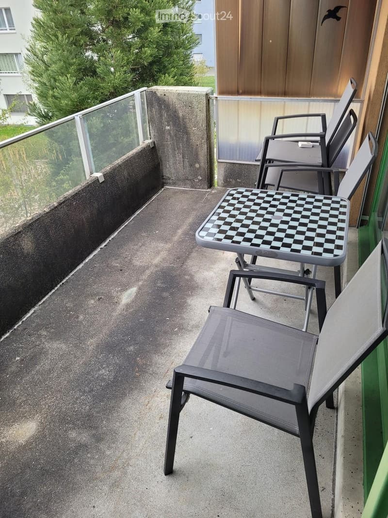 Balcony with a concrete floor, glass railing, and outdoor furniture including a checkered table and several chairs.