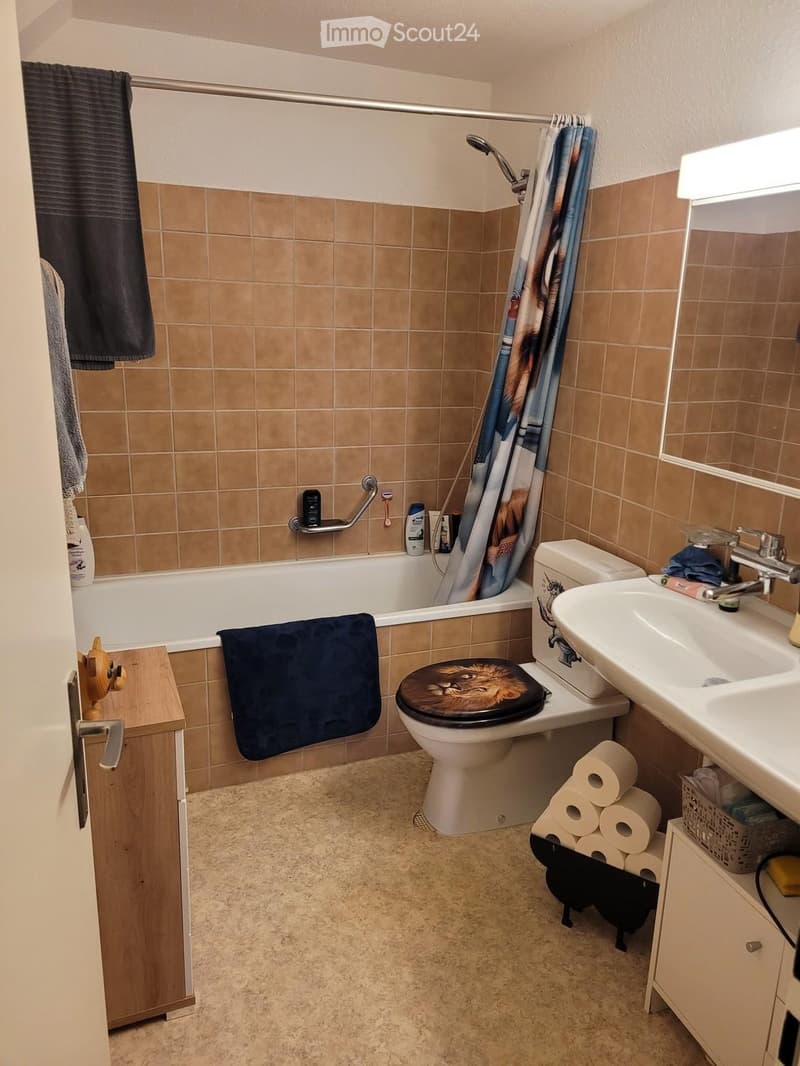 Bathroom with beige tiles, bathtub, toilet, sink, mirror, towel, toilet paper, and a shower curtain.