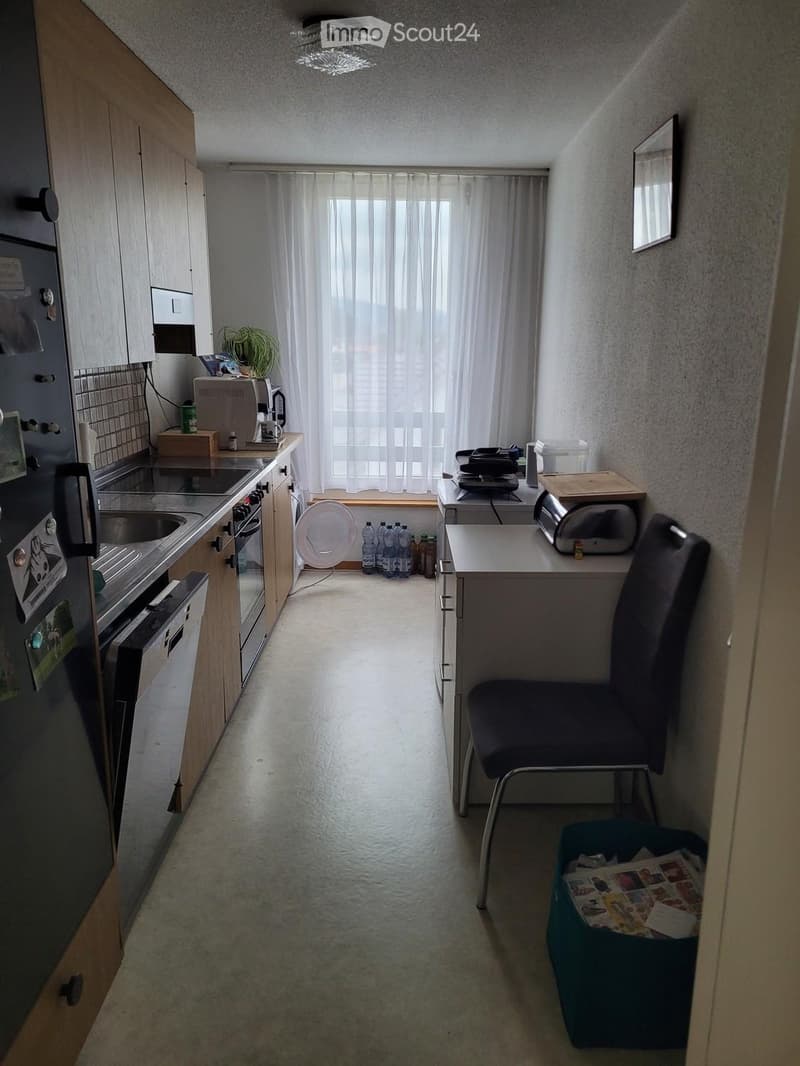 Compact kitchen with wooden cabinets, stainless steel appliances, a small table with a chair, and a window with white curtains.