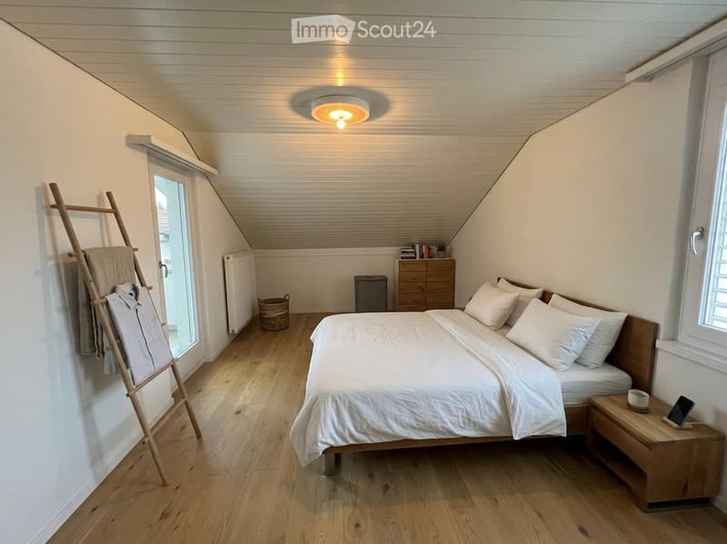 Bright, modern bedroom with wooden flooring, slanted ceiling, and large windows. Features a double bed with white bedding, a wooden nightstand with a lamp, a small dresser, and a coat rack with folded clothes.