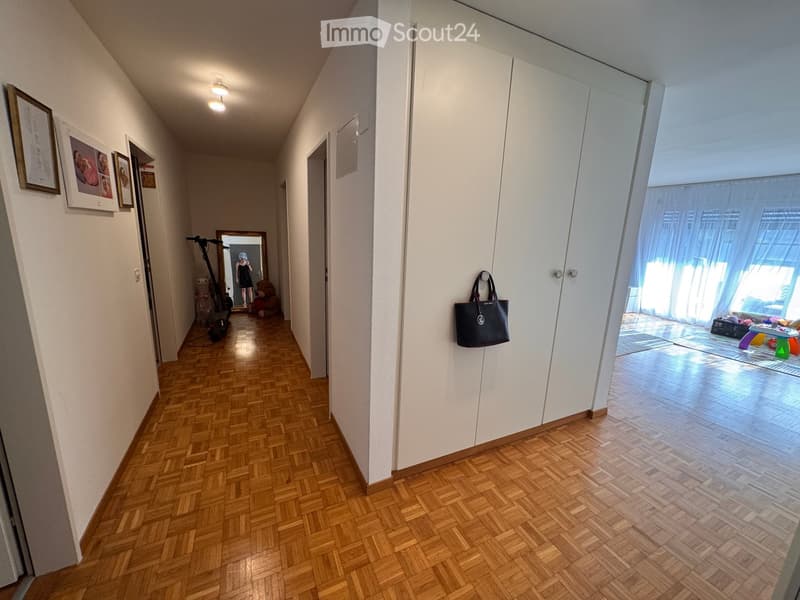 A well-lit hallway with wooden herringbone flooring, white walls, and several doors leading to different rooms. A large white closet is visible on the right, and a black handbag is hanging on it. The hallway features framed pictures on the left wall and a