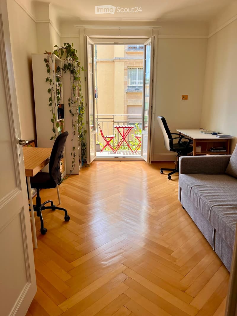 A modern, well-lit living space with wooden flooring, a grey sofa, a desk, and a balcony with outdoor furniture.