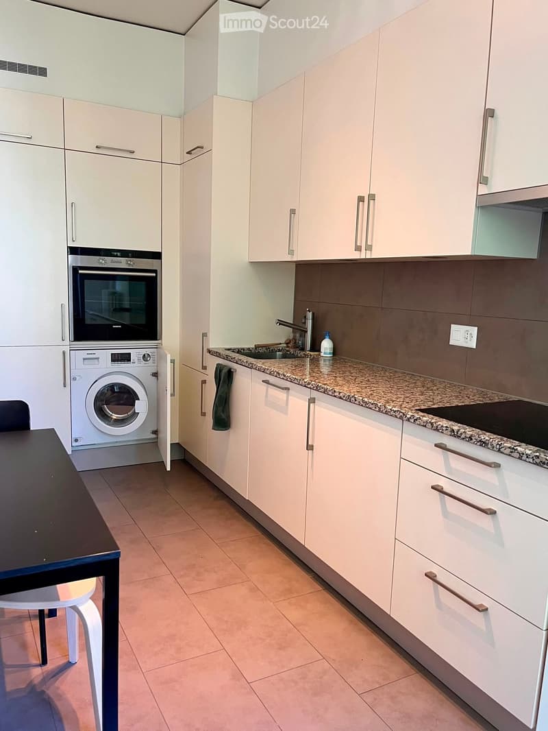 Modern kitchen with white cabinets, granite countertop, built-in oven, washing machine, and a small dining area with a table and chair.