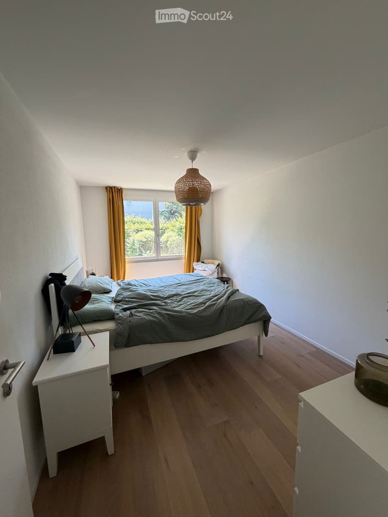 A cozy bedroom with a double bed, nightstands, a hanging lamp, and a window with curtains. The room features wooden flooring and white walls.