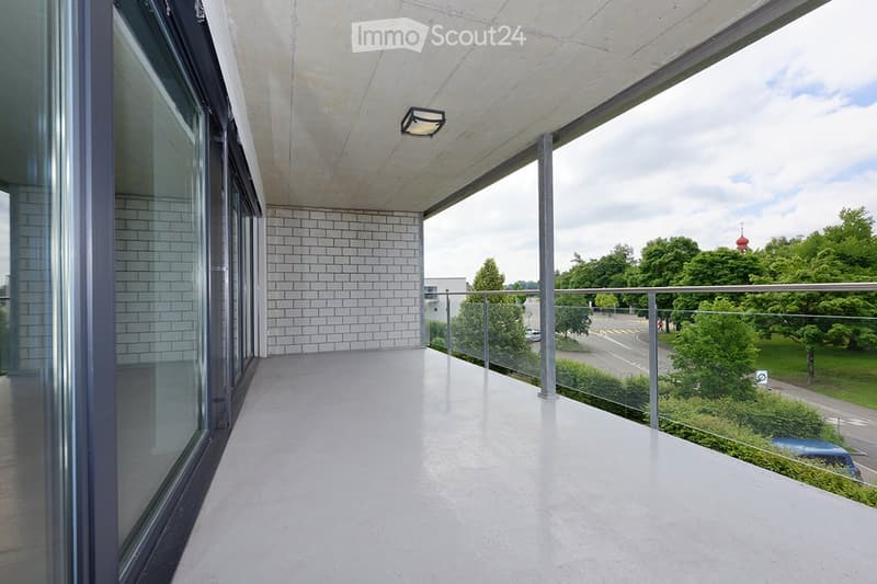 Modern balcony with large windows, white tiled walls, and a view of greenery and a road.