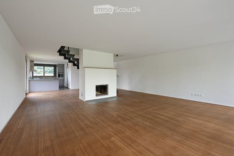 Open-plan living space with wooden flooring, white walls, and a central fireplace. The area includes a staircase leading to an upper level and a visible kitchen area with cabinets and a window.