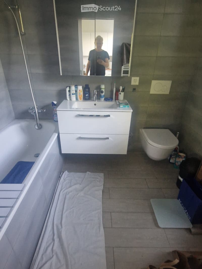 Modern bathroom with a white sink, mirror, toilet, bathtub, and various toiletries on the sink counter. A towel is placed on the floor.