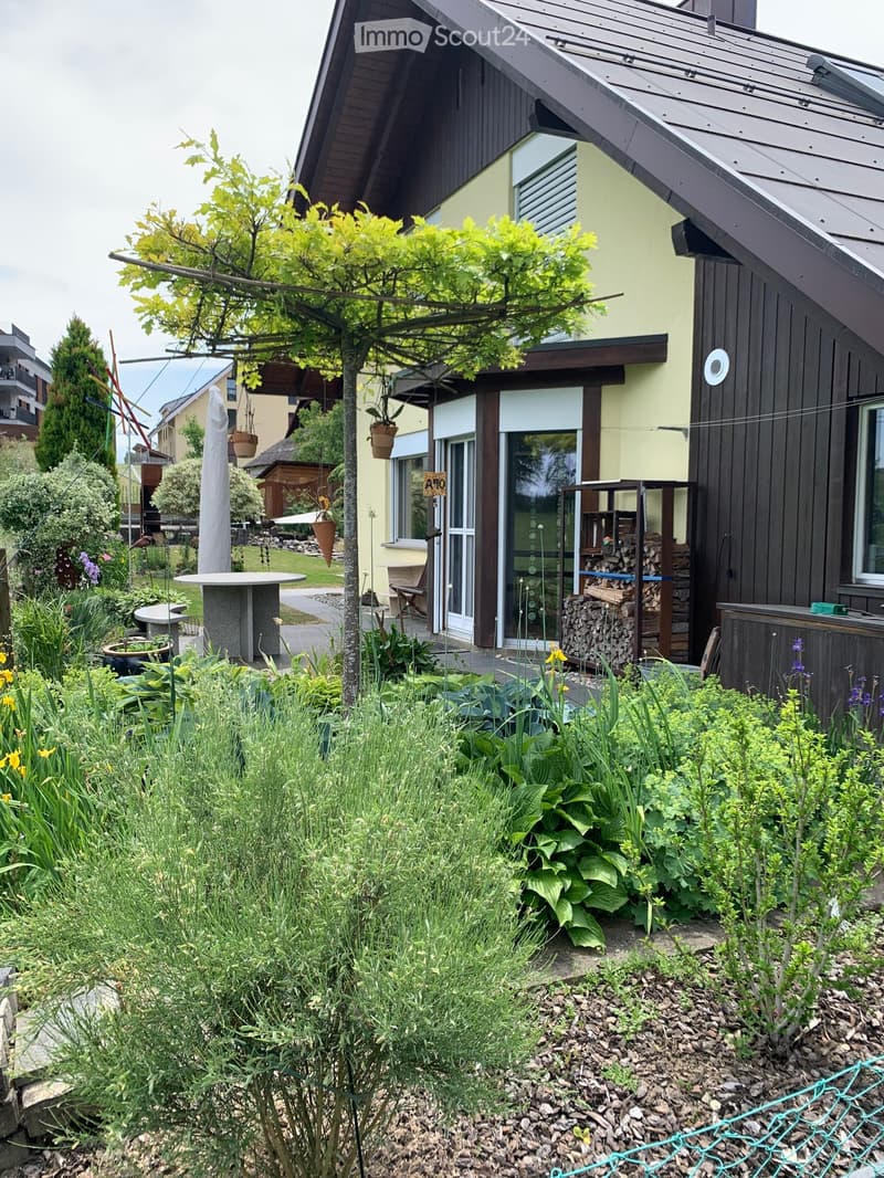 Garden area with a modern house in the background, featuring a variety of plants, a round table, a stack of firewood, and a hanging basket.