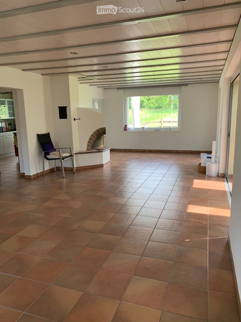 Spacious room with terracotta tiled floor, fireplace, and a single chair. Large window with view of greenery.