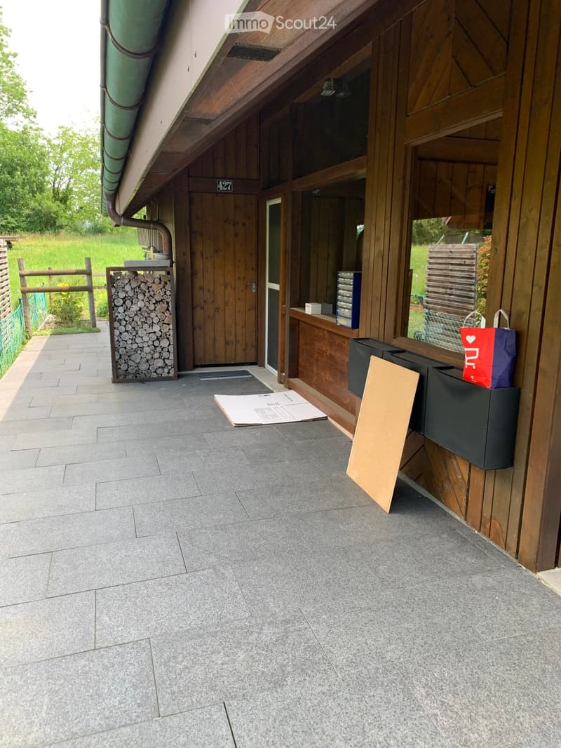 Balcony with grey tiled floor, wooden walls, and a stack of firewood to the left. A mailbox and a shopping bag are visible on the right side.