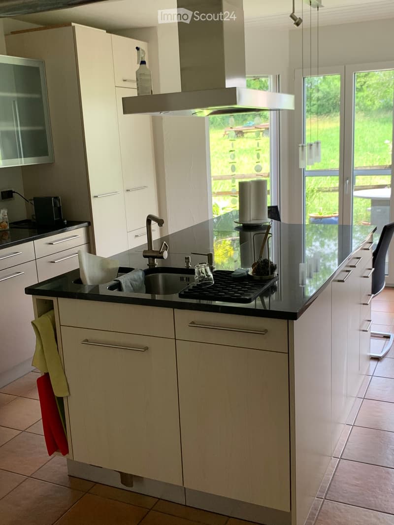 Modern kitchen with white cabinets, black countertops, a sink, and a stove. There is a chair near the counter and a view of the garden through large windows.