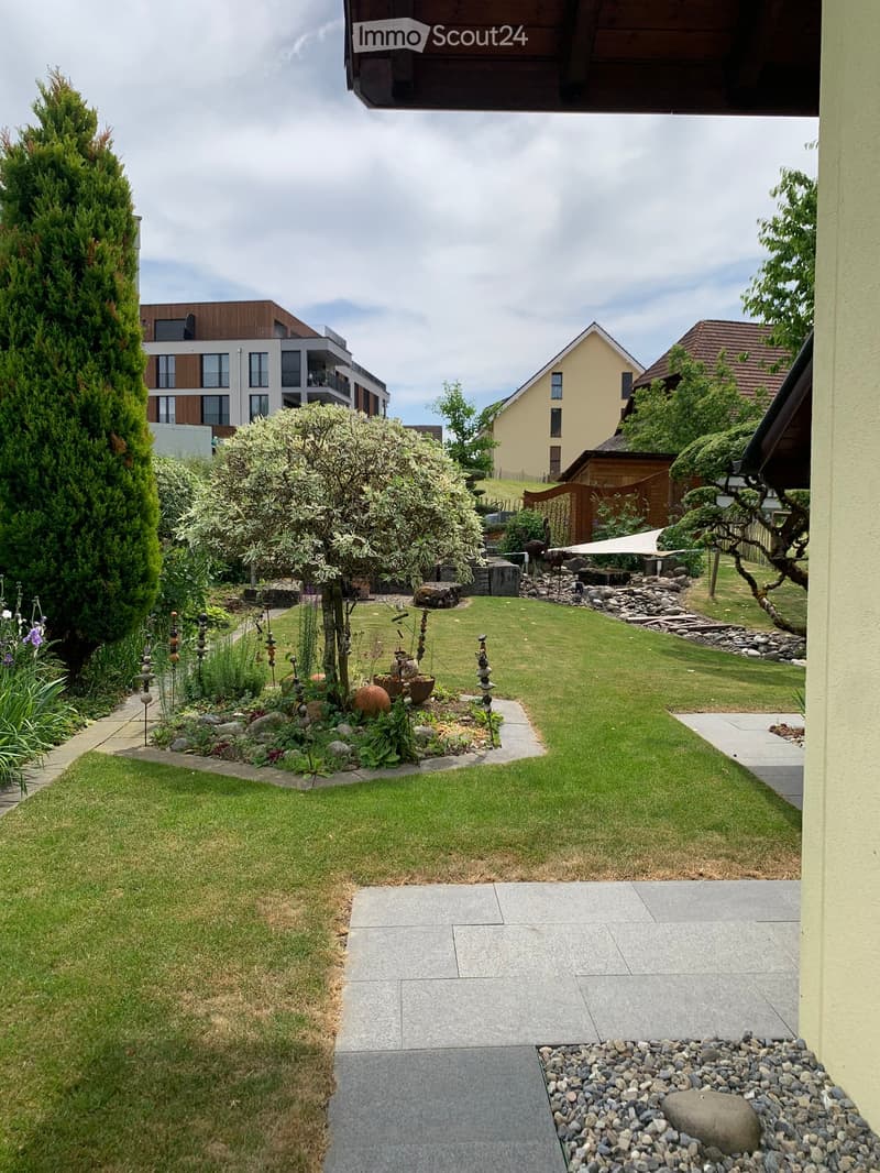 Well-maintained garden with a lawn, stone pathways, decorative rocks, and surrounding trees and shrubs.