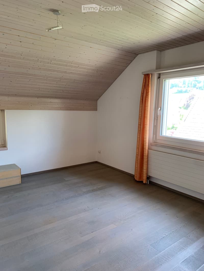Empty room with wooden floor, white walls, slanted wooden ceiling, window with curtain, and a small wooden chest.