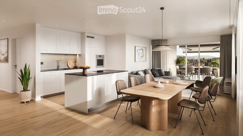 Open-plan living and dining area with modern white kitchen cabinets, a central island, a large wooden dining table with chairs, and a grey sofa. The space features a pendant light, artwork, and a potted plant, with large windows offering a view of the out