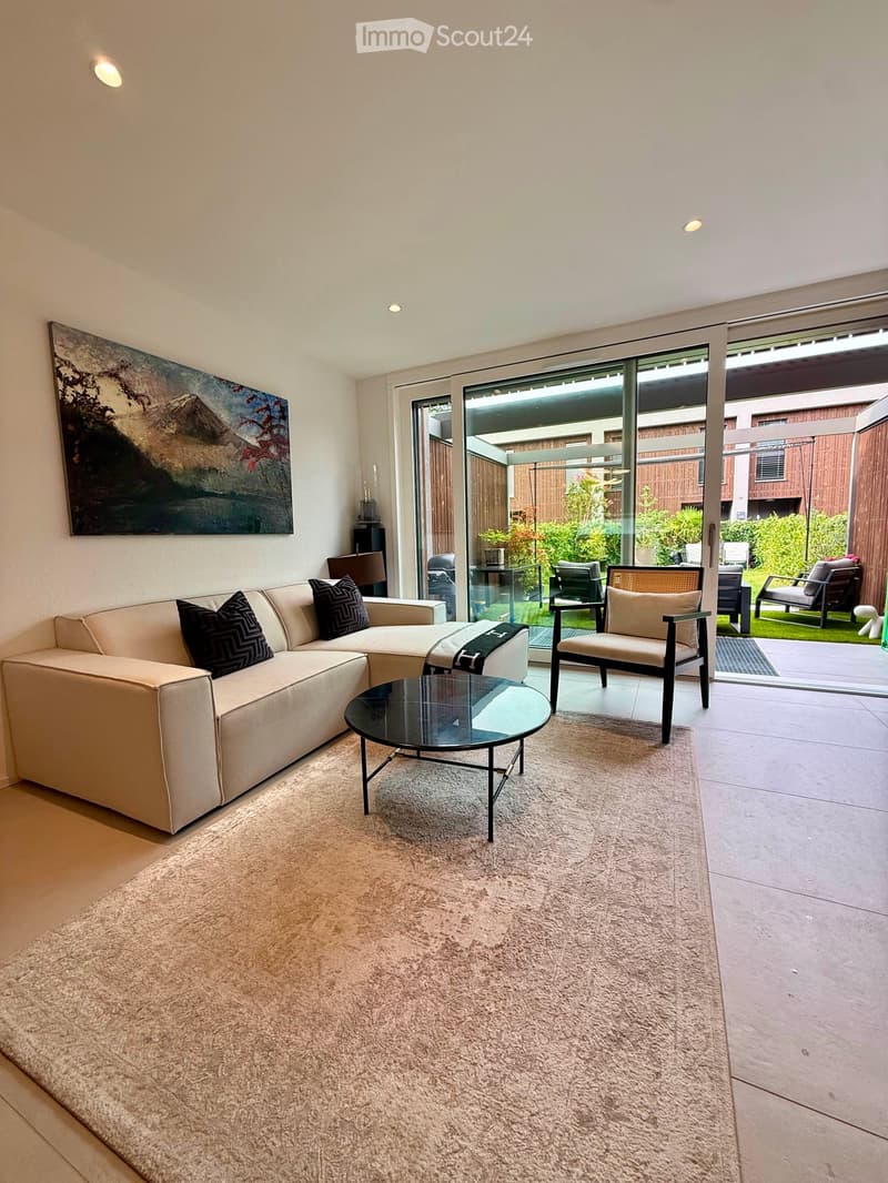 Modern living room with a beige sectional sofa, black throw pillows, a glass coffee table, a large area rug, and a painting on the wall. Large glass doors open to a patio with outdoor seating.