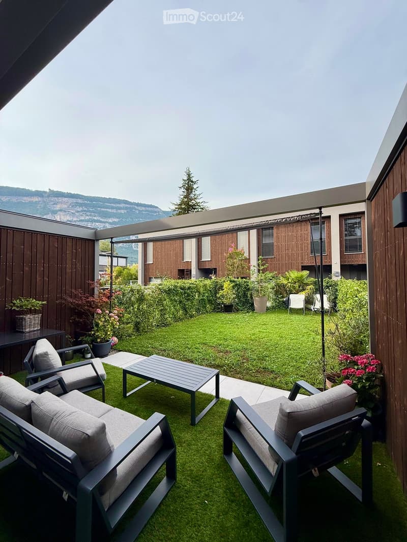 A modern outdoor patio with artificial grass, grey furniture including a sofa, chairs, and a coffee table, surrounded by potted plants and a view of a landscaped garden and mountains.