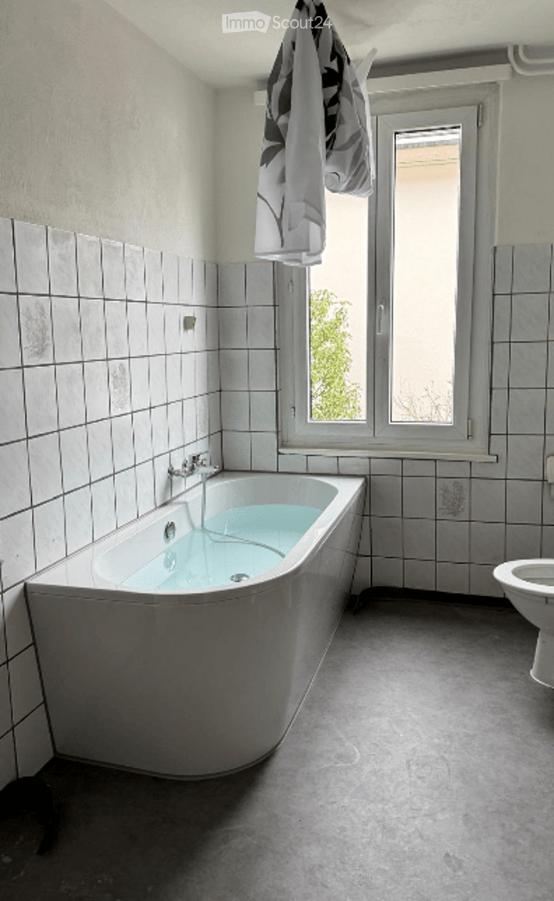 Bathroom with a white bathtub, toilet, and tiled walls. A towel hangs on a rack near the window, which offers a view of greenery outside.