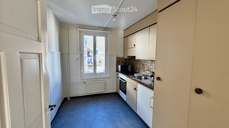 Compact kitchen with white cabinets, a sink, microwave, and oven. The space features a window with a view of the outside and a radiator below it.