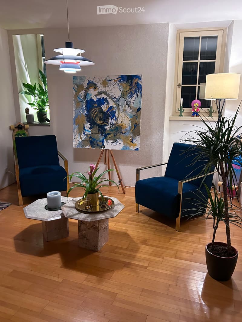 Modern living room with wooden flooring, two blue armchairs, a marble coffee table, a floor lamp, and a painting on the wall.