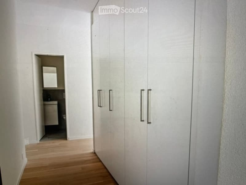 A narrow, well-lit hallway with light wooden flooring and white walls. A set of white built-in wardrobes with silver handles lines one side, and an open doorway leads to a small bathroom with a sink and mirror.
