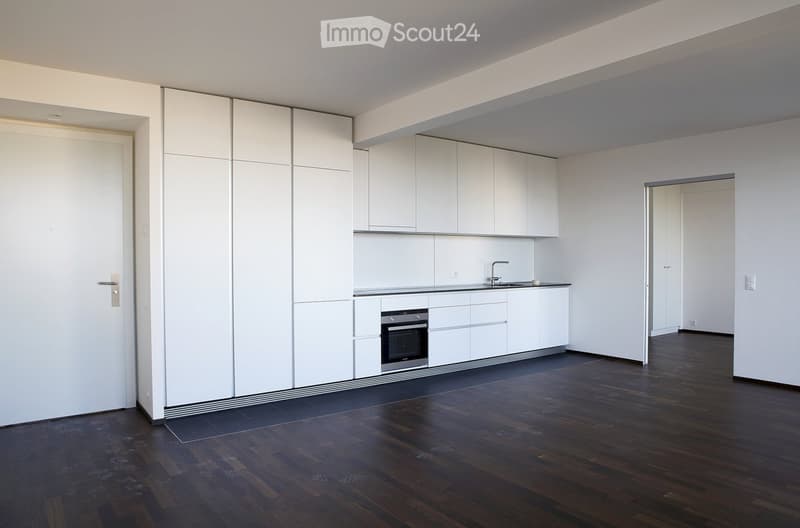 Empty room with white walls, dark wooden flooring, and a small white kitchen area featuring cabinets, a sink, and an oven. The space is clean and unfurnished.