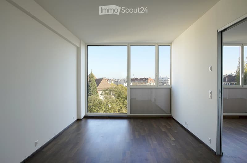 Empty room with wooden flooring, white walls, large windows offering a view of the city and trees, and modern electrical outlets.