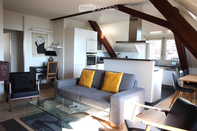 Modern living room with exposed wooden beams, grey sofa with yellow cushions, glass coffee table, dining area with table and chairs, and an open kitchen with white cabinets and stainless steel appliances.