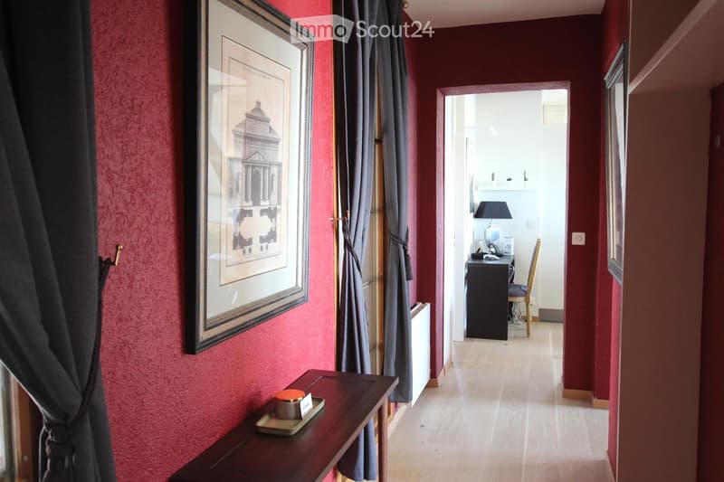 A narrow hallway with red walls, wooden flooring, and a framed picture on the wall. A small table with a decorative item sits near the window, and an open doorway leads to another room with a desk, chair, and lamp.