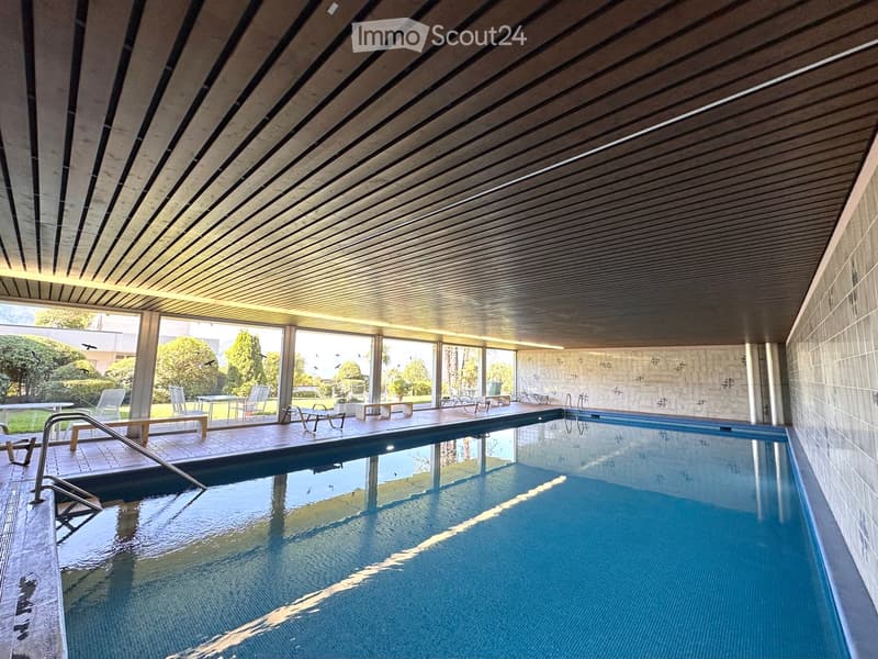 Indoor pool area with large windows, surrounded by lounge chairs and tables, with a view of the outdoor garden and trees.