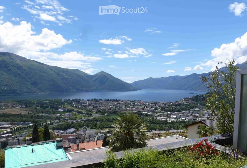 Panoramic view from a balcony or terrace overlooking a town, lake, and surrounding mountains under a partly cloudy sky.