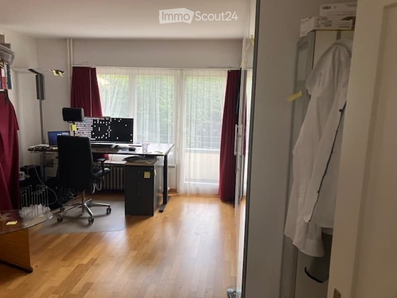 A furnished room with a wooden floor, large window with curtains, a desk with a computer and screen, a chair, and a lamp.
