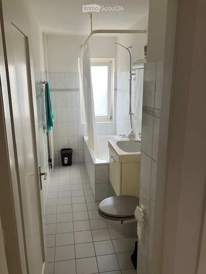 A small, clean bathroom with white tiled walls and floor, featuring a bathtub, sink, toilet, and a shower area with a glass door. A towel hangs on a rack, and a trash bin is placed near the door.