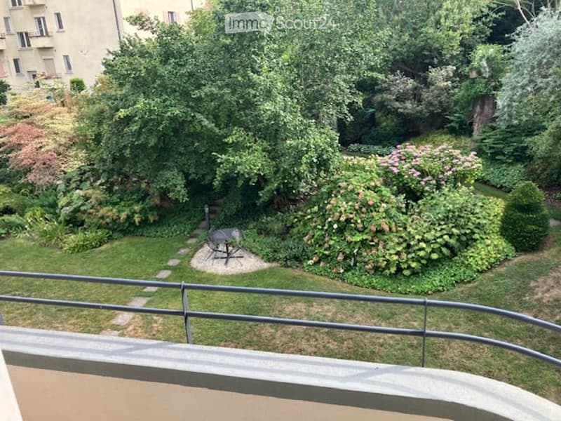 Garden view from a balcony, featuring a well-maintained lawn, various trees and shrubs, a small seating area with a table and chairs, and a stone pathway.