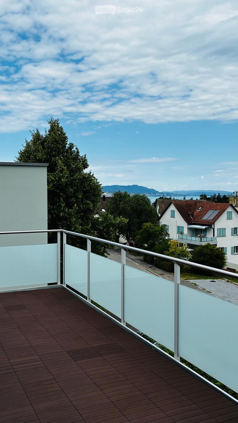 Balcony with wooden decking, glass railing, overlooking a suburban area with trees and houses.